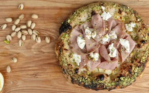 Pizza mit Mortadella, Pistazien und cremigem Käse, daneben einige Pistazien und eine Zitronenspalte.