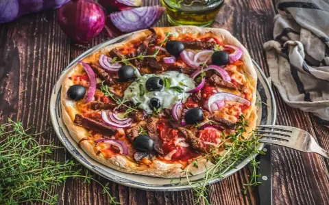 Herzhafte Gyros Pizza mit Zwiebeln, Oliven und Kräutern, serviert auf einem Teller mit Gabel und Messer.