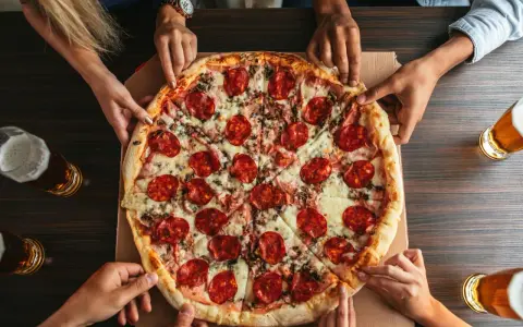 Hände greifen nach einer großen Salamipizza, die auf einem Tisch liegt. Jeder bekommt seinen fairen Anteil an pizzateig pro person.