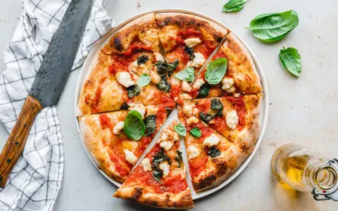 Eine köstliche, frisch gebackene pizza vegana mit Tomaten, Basilikum und veganem Mozzarella. Daneben ein Messer und ein Glas Öl.