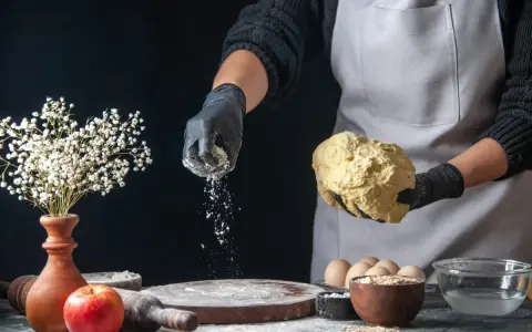 Hände in schwarzen Handschuhen streuen selbsttreibendes Mehl über Teig. Ein Apfel und Blumen sind auf dem Tisch.