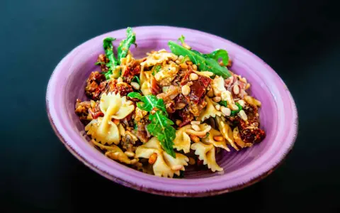 Ein köstlicher italienischer Nudelsalat mit Farfalle, sonnengetrockneten Tomaten, Pinienkernen und Rucola in einer lila Schüssel.