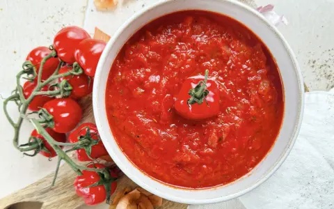 Eine Schüssel mit köstlicher, hausgemachter italienischer Tomatensauce, garniert mit einer frischen Kirschtomate. Daneben liegen frische Tomaten und Zwiebeln.