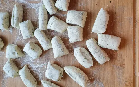 Vorbereitung für ein Grissini Rezept: Viele kleine, unregelmäßig geformte Teiglinge mit Kräutern liegen auf einem Holzbrett, bestäubt mit Mehl.
