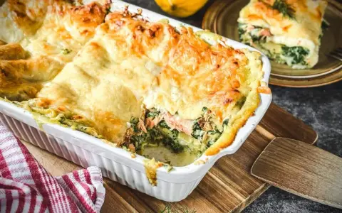 Herzhafte Lachs Spinat Lasagne, goldbraun überbacken, dampft in einer weißen Auflaufform. Ein Stück auf einem Teller daneben.