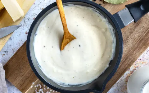 Cremige Bechamelsauce in einer Pfanne, bereit für die Lasagne. Ein Holzlöffel rührt die sämige Masse.