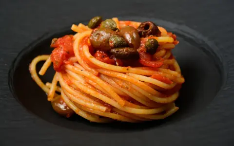 Ein kunstvoll aufgetürmter Teller Spaghetti Puttanesca, mit Oliven und Kapern garniert, auf schwarzem Teller.