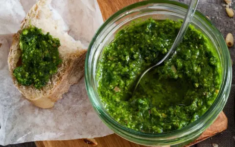 Frisches schnittlauch pesto in einem Glas, mit einem Löffel darin. Ein Stück Brot mit dem grünen Pesto darauf liegt daneben.