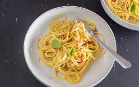 Ein Teller mit cremiger Spaghetti Carbonara, garniert mit Basilikum und Speckstückchen.