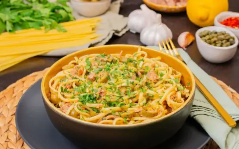 Eine Schüssel mit köstlicher Thunfisch Pasta, garniert mit Petersilie und Zitronenschale. Daneben liegen Spaghetti, Knoblauch und Kapern.