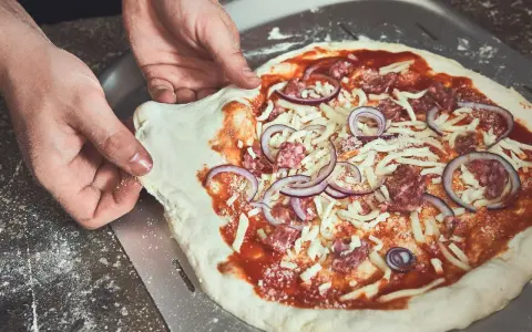 Hände schieben einen rohen Pizzaboden mit Tomatensauce, Käse, Salami und Zwiebeln auf einen Pizzaschieber. Der pizzateig klebt am schieber.