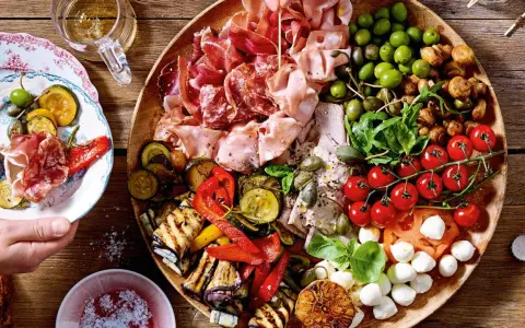 Eine üppige italienische Antipasti-Platte wird liebevoll dekoriert: Wurst, Käse, Oliven, gegrilltes Gemüse und frische Kräuter laden zum Genuss ein.