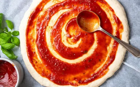 Eine Pizza nur mit Tomatensauce wird mit einer Löffelspirale belegt. Frisches Basilikum liegt daneben.