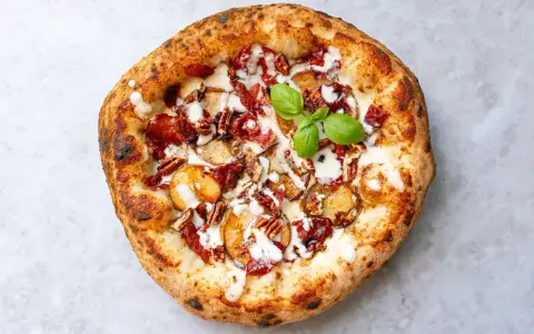 Pizza mit karamellisierten Zwiebeln, Speck, Pekannüssen und einem Hauch von Parmesan. Ein grünes Basilikumblatt krönt das Kunstwerk.