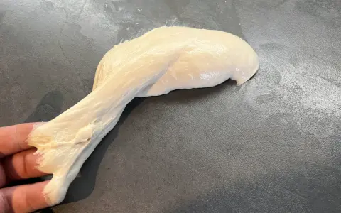 Ein Stück heller, klebriger Pizzateig wird von einer Hand gehalten und dehnt sich auf einer dunklen Arbeitsfläche aus.
