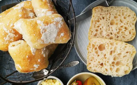 Frisch gebackene Ciabatta-Brötchen, bestäubt mit Mehl, liegen in einem Korb. Daneben zwei aufgeschnittene Scheiben mit luftiger Krume – perfekt zum ciabatta selber backen.