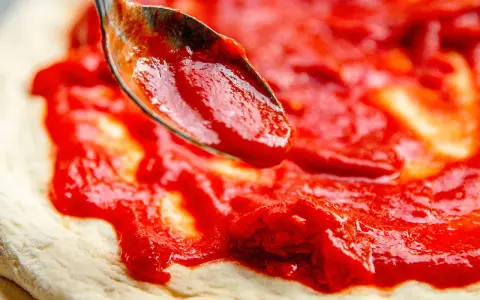 Eine Löffelspitze verteilt die leuchtend rote Tomatensauce auf dem Teig für eine authentische Pizza Napoletana.