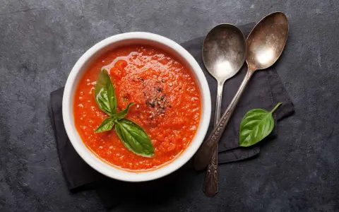 Schüssel mit selbstgemachter Tomatensauce, garniert mit Basilikum und Pfeffer, daneben zwei Löffel auf dunklem Hintergrund.