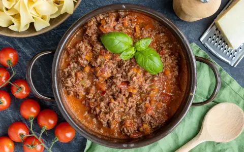 Herzhaftes Bolognese Rezept in einem Topf, garniert mit Basilikum. Daneben frische Tomaten, Bandnudeln und Parmesan.