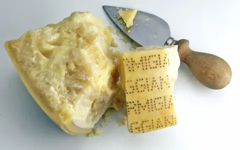 Ein Stück Parmesan mit eingeprägtem Schriftzug und ein Käsemesser. Aus was besteht Parmesan? Aus Milch, Lab und Salz.