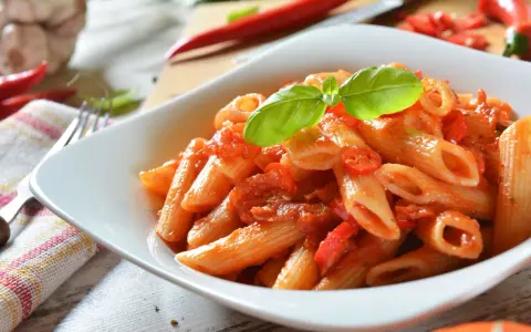 Eine Schüssel Penne Arrabiata, garniert mit Basilikum, mit scharfen Chilischoten und Knoblauch im Hintergrund.
