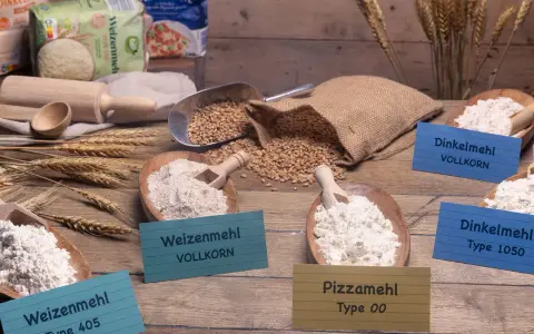 Verschiedene Mehlsorten, darunter Weizenmehl, Dinkelmehl und Pizzamehl, liegen in Holzschalen. Daneben Weizenkörner und ein Nudelholz.