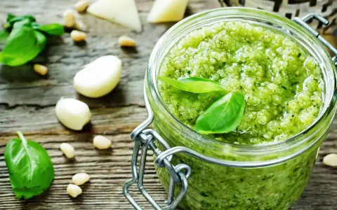 Frisches Pesto-Rezept im Glas, umgeben von Basilikum, Pinienkernen, Knoblauch und Parmesan auf rustikalem Holz.