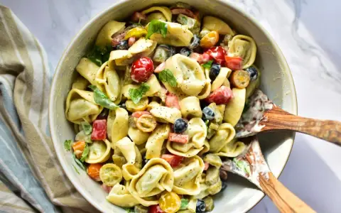 Ein cremiger Tortellini Salat mit Kirschtomaten, Oliven und Kräutern, serviert in einer Schüssel mit Holzspateln.