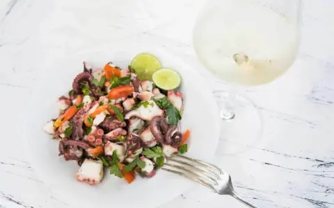 Frischer Insalata di polpo mit Tomaten, Petersilie und Limettenspalten auf einem Teller, daneben ein Glas Weißwein.