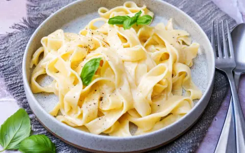 Cremige Pasta Alfredo mit frischem Basilikum und schwarzem Pfeffer, serviert auf einem grauen Tischtuch.