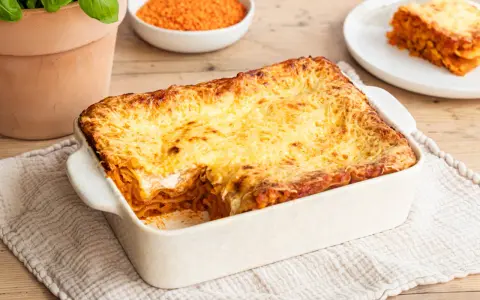 Herzhafte Linsen Lasagne mit goldbraun überbackener Käseschicht, daneben eine Schüssel roter Linsen und ein Teller mit einem Stück Lasagne.