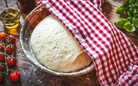 Frischer Pizzateig ruht in einer Schüssel, bedeckt mit einem rot-weiß karierten Tuch. Die Frage: wie viel Hefe in Pizzateig?