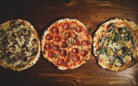 Drei leckere Pizza-Sorten: eine mit Pilzen, eine mit Salami und eine mit Gemüse, liegen nebeneinander auf einem Holztisch.