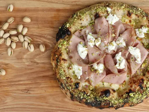 Pizza mit Mortadella, Pistazien und cremigem Käse, daneben einige Pistazien und eine Zitronenspalte.
