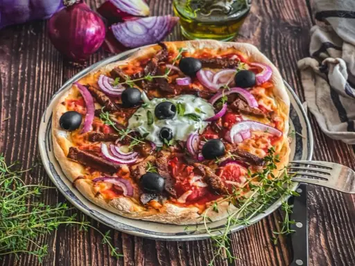Herzhafte Gyros Pizza mit Zwiebeln, Oliven und Kräutern, serviert auf einem Teller mit Gabel und Messer.
