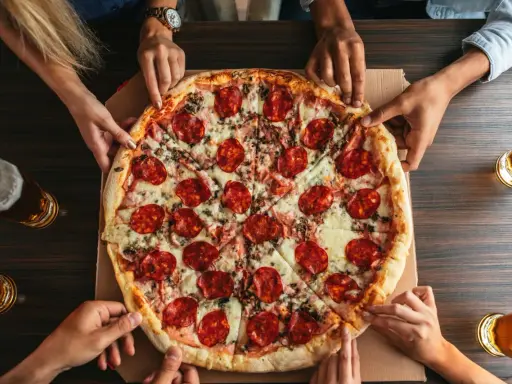 Hände greifen nach einer großen Salamipizza, die auf einem Tisch liegt. Jeder bekommt seinen fairen Anteil an pizzateig pro person.