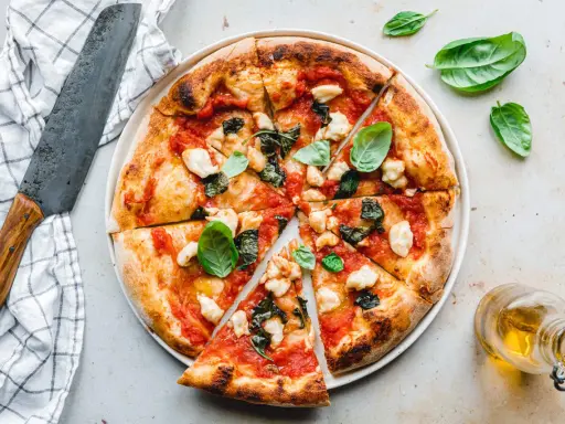 Eine köstliche, frisch gebackene pizza vegana mit Tomaten, Basilikum und veganem Mozzarella. Daneben ein Messer und ein Glas Öl.