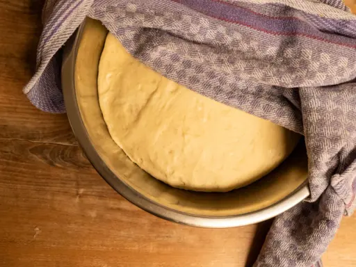 Frischer, aufgegangener Pizzateig ruht in einer Schüssel, bedeckt mit einem gemusterten Tuch. Perfekt für Ihr Pizzateig Rezept.