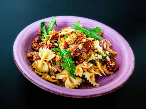 Ein köstlicher italienischer Nudelsalat mit Farfalle, sonnengetrockneten Tomaten, Pinienkernen und Rucola in einer lila Schüssel.