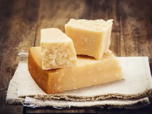 Drei Stücke Hartkäse, vielleicht Pecorino oder Parmesan, liegen auf einem Leinentuch vor rustikalem Holz.