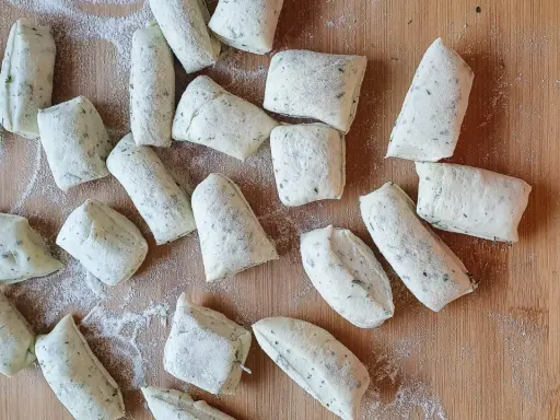 Vorbereitung für ein Grissini Rezept: Viele kleine, unregelmäßig geformte Teiglinge mit Kräutern liegen auf einem Holzbrett, bestäubt mit Mehl.