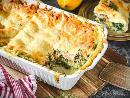 Herzhafte Lachs Spinat Lasagne, goldbraun überbacken, dampft in einer weißen Auflaufform. Ein Stück auf einem Teller daneben.