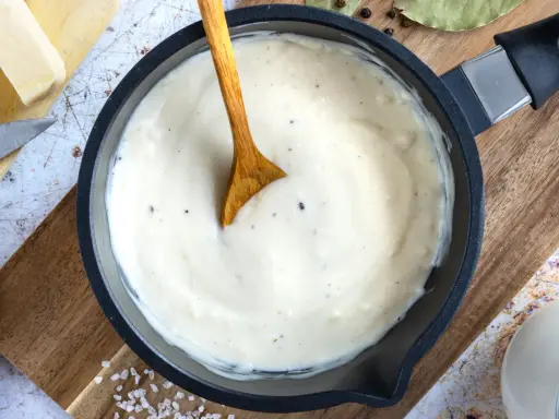 Cremige Bechamelsauce in einer Pfanne, bereit für die Lasagne. Ein Holzlöffel rührt die sämige Masse.