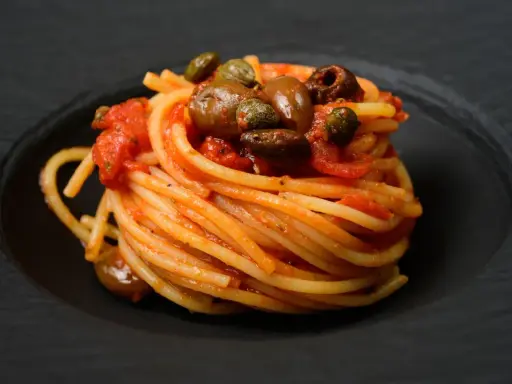 Ein kunstvoll aufgetürmter Teller Spaghetti Puttanesca, mit Oliven und Kapern garniert, auf schwarzem Teller.