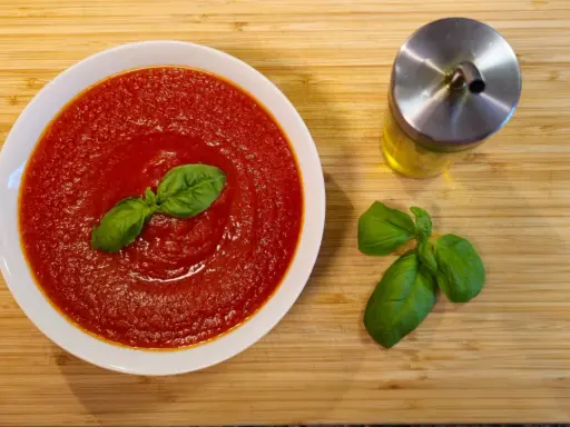 Schüssel mit cremiger Tomatensauce, garniert mit Basilikum. Daneben ein Glas Olivenöl und frische Basilikumblätter. Perfekt für Pasta mit San Marzano Tomaten.