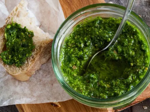 Frisches schnittlauch pesto in einem Glas, mit einem Löffel darin. Ein Stück Brot mit dem grünen Pesto darauf liegt daneben.