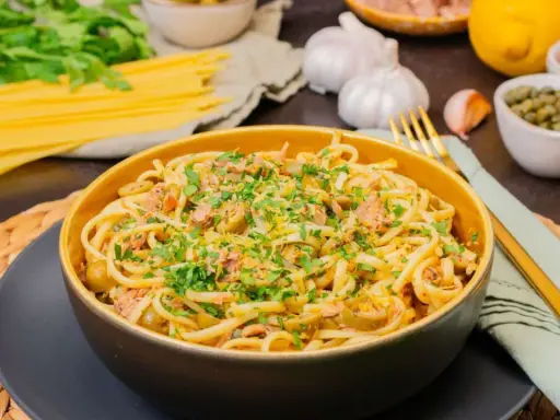 Eine Schüssel mit köstlicher Thunfisch Pasta, garniert mit Petersilie und Zitronenschale. Daneben liegen Spaghetti, Knoblauch und Kapern.