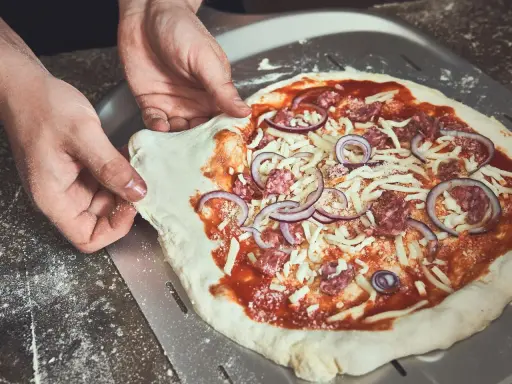Hände schieben einen rohen Pizzaboden mit Tomatensauce, Käse, Salami und Zwiebeln auf einen Pizzaschieber. Der pizzateig klebt am schieber.