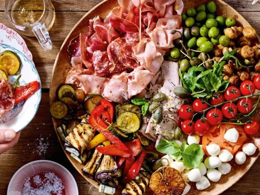 Eine üppige italienische Antipasti-Platte wird liebevoll dekoriert: Wurst, Käse, Oliven, gegrilltes Gemüse und frische Kräuter laden zum Genuss ein.
