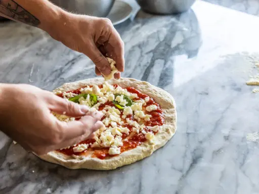 Hände belegen Pizza mit zerbröckeltem Käse, einer köstlichen fior di latte Alternative, auf Marmortisch.